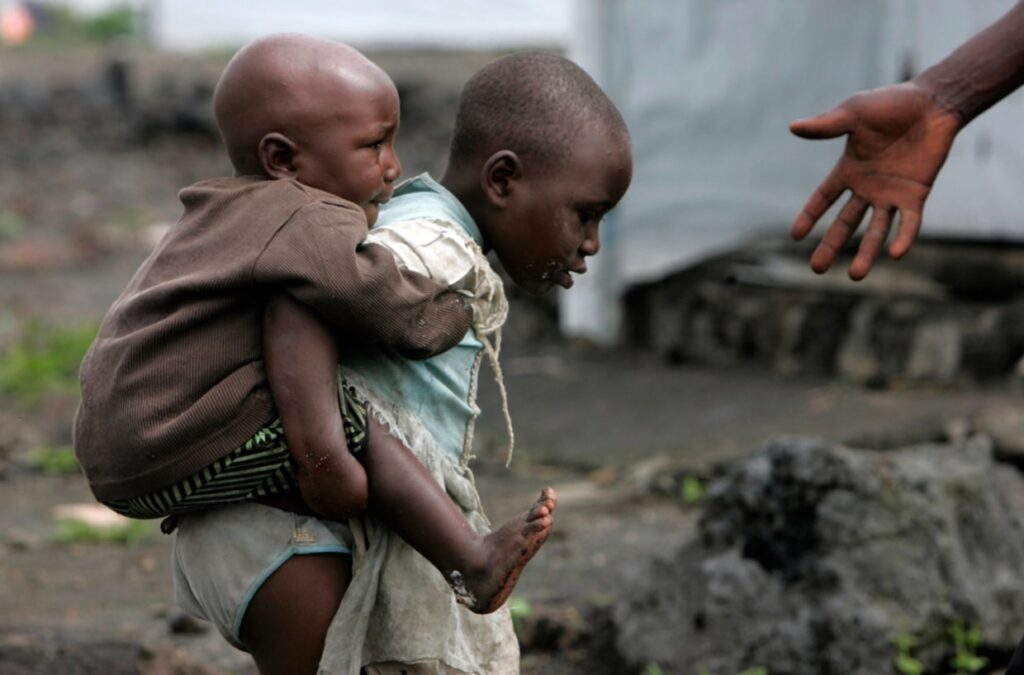 A man reaches to help two displaced children cross sharp lava rock in a camp for displaced people in Kibati just north of Goma in eastern Congo. Photo Credit :Karel Prinsloo / AP